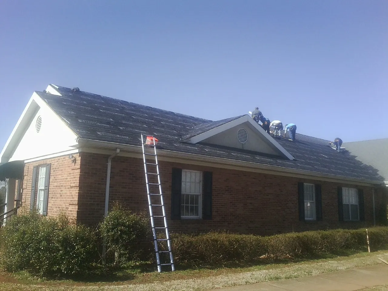 Crew tearing shingles off a brick ranch roof with a ladder set — Metal Roof Repair work by our crew in Byron Center