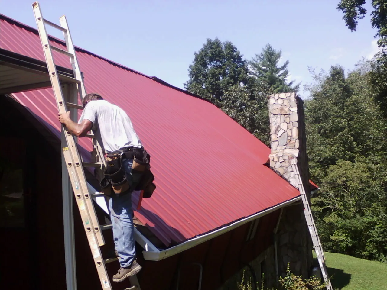 Roofer on a ladder beside a red metal roof and stone chimney — Soffit & Fascia Repair by Roofing Brothers in Byron Center